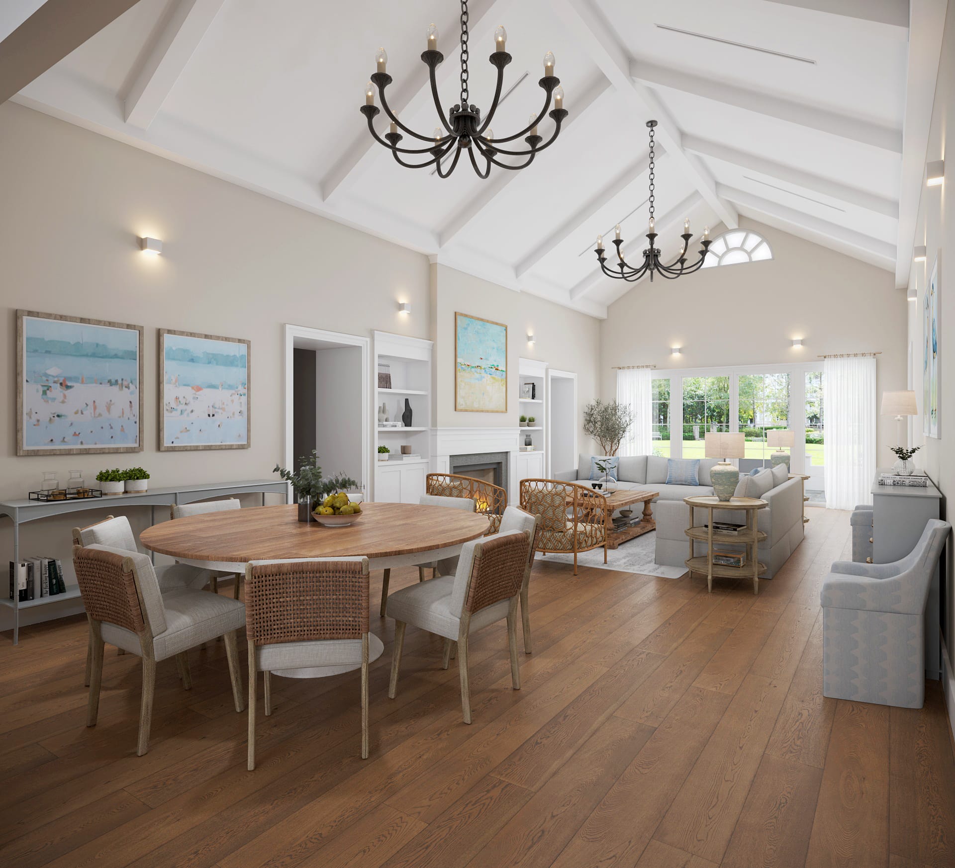 Open-plan coastal great room with a round oak dining table and woven chairs in the foreground, vaulted ceiling with painted rafters, two iron chandeliers and a grey sectional by a glazed wall beyond